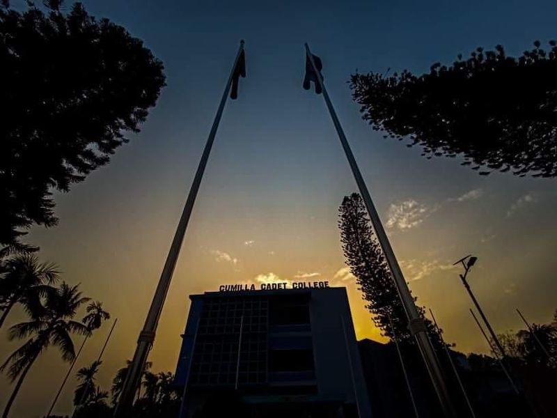 Two flagpoles silhouetted above Cumilla Cadet College at sunset